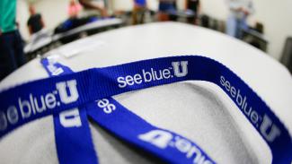 A lanyard with the see blue. U printed down the side lays on a table in front of an out of focus room of people.